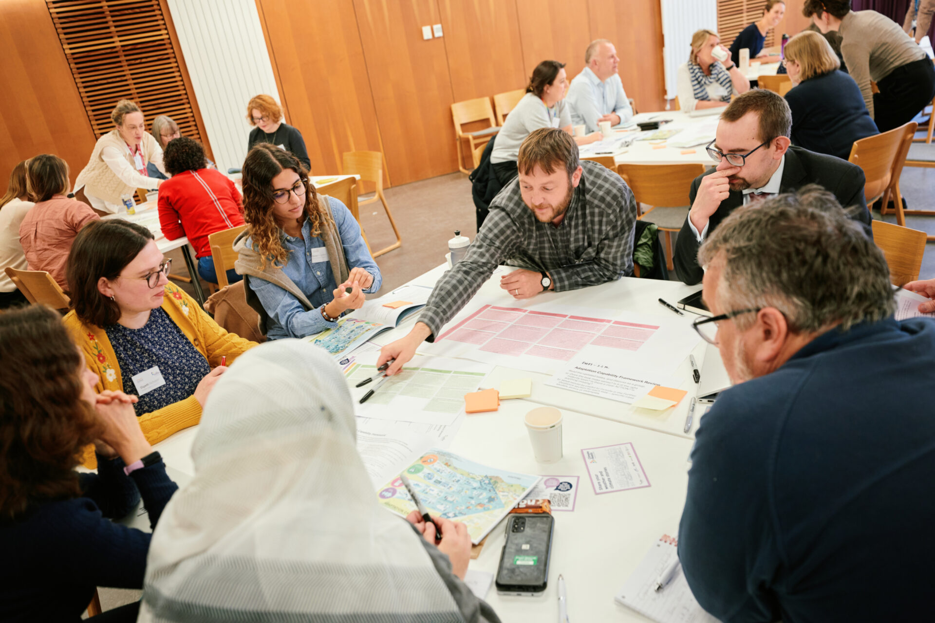Photo showing 6 people sat around a table with papers, pens, and post-it notes. One person points to one of the papers, leaning over. Other people sat at tables are visible in the background